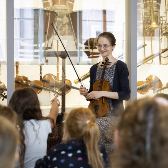 Kobieta trzymająca skrzypce i grupa dzieci na tle gabloty z instrumentami