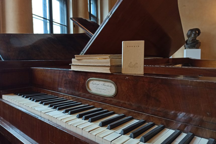 Kilka książek ułożonych na fortepianie, w tym tomik poezji C.K. Norwida. W tle widoczne popiersie F. Chopina.