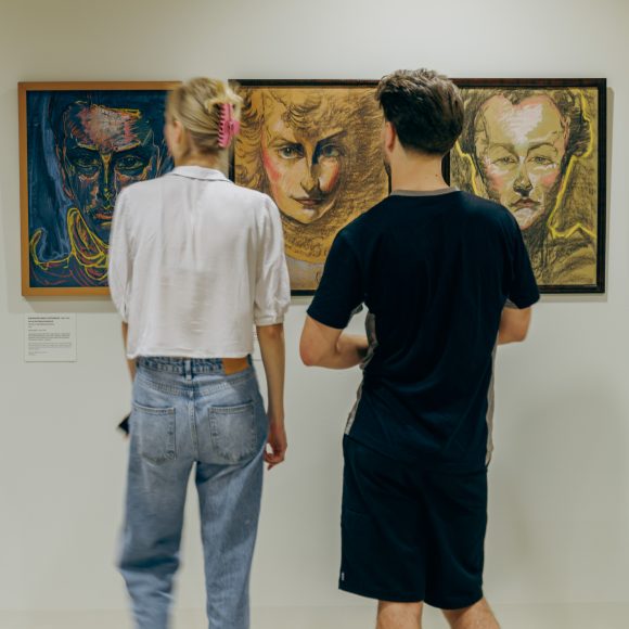 Dwie osoby stojące tyłem patrzą na ścianę, na której wiszą trzy obrazy.