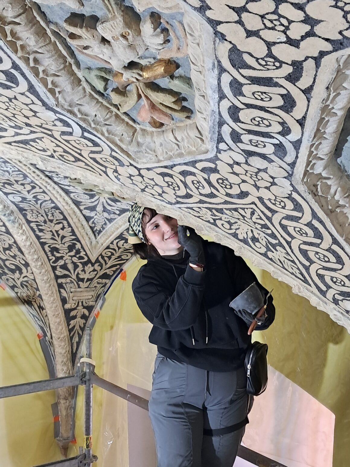 1 Kobieta ubrana w czarna bluzę i robocze spodnie i czarne rękawiczki dotyka dłonią w rękawiczce fragment zabytkowego sufitu.