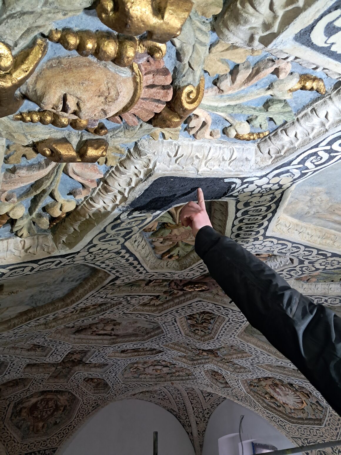 3 Fragment dekoracji zabytkowego sufitu. Od prawej strony ludzka ręka wskazująca palcem wskazującym środek dekoracji.