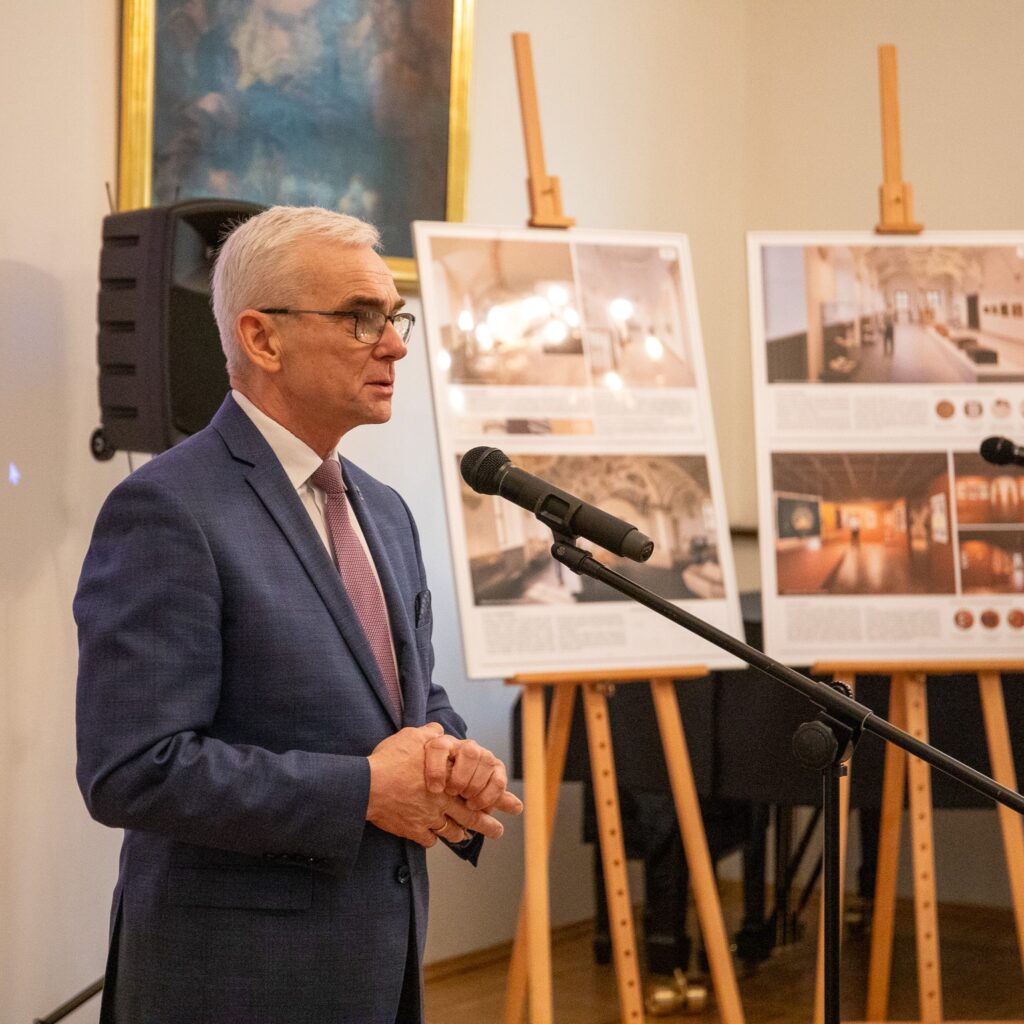 Mężczyzna przy mikrofonie. W tle sztalugi z planszami.