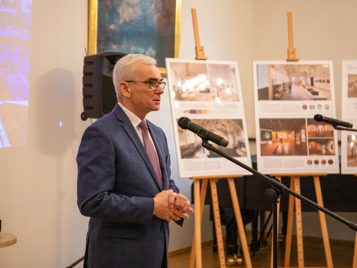 _MG_1633 Mężczyzna stojący przy mikrofonie. W tle sztalugi z planszami.