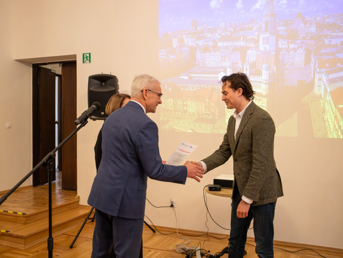 _MG_1692 Dwóch mężczyzn ściska sobie dłonie.W tle jasną ściana z rzuconym przez rzutnik zdjęciem miasta.