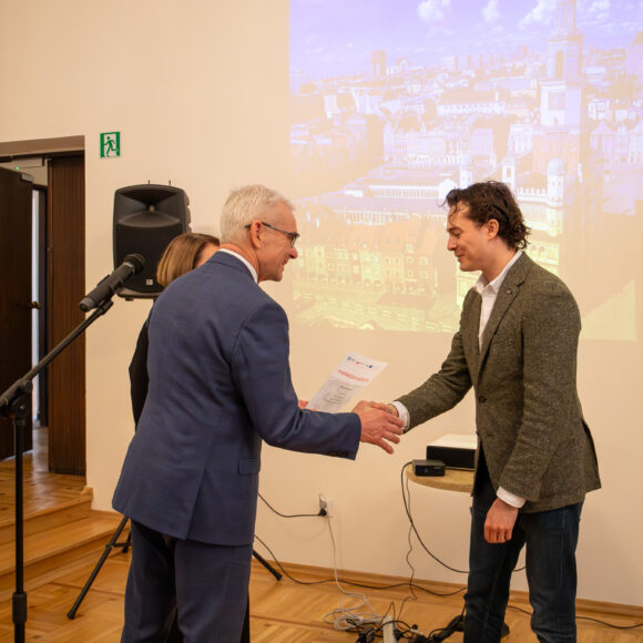 _MG_1692 Dwóch mężczyzn ściska sobie dłonie.W tle jasną ściana z rzuconym przez rzutnik zdjęciem miasta.