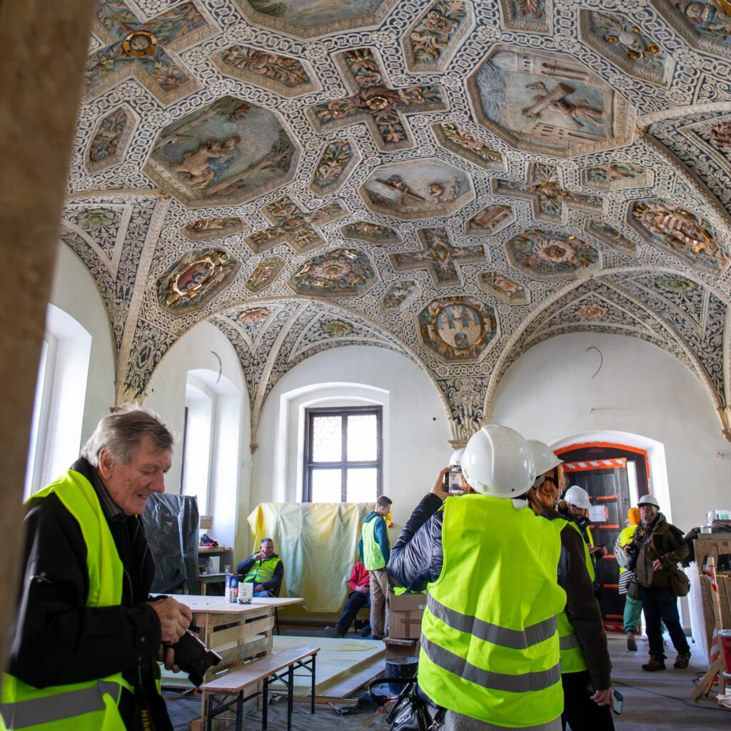 Kilka osób w białych kaskach i odblaskowych kamizelkach w zabytkowym pomieszczeniu.