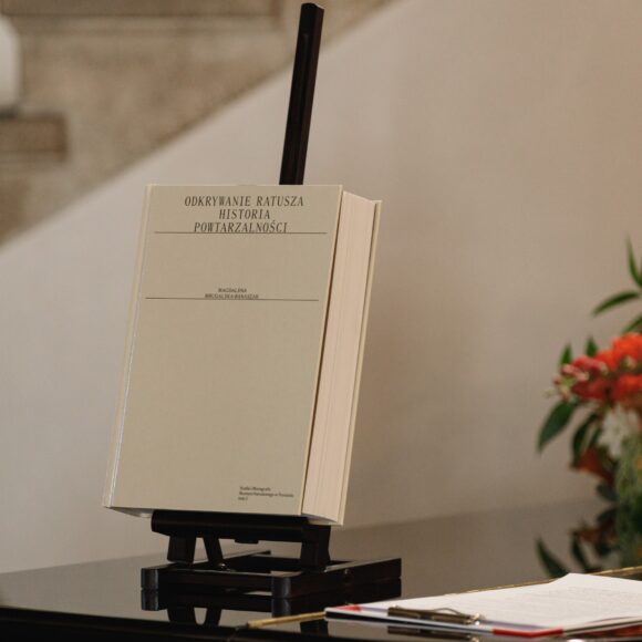 2025.05.07_Premiera_książki_dr.Magdaleny_Mrugalska_Banaszak_ratusz-24 Książka z białą okładką ustawiona na małej czarnej sztaludze.