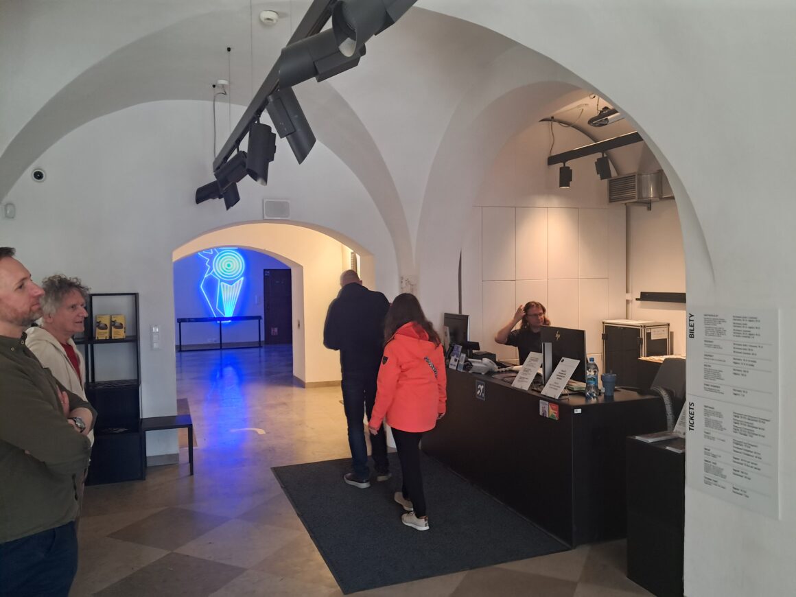 Fragment pomieszczenia z czarną ladą. Z boku tablica z cennikiem biletów. Za ladą siedzi jeden pracownik. W części przed ladą znajdują śsię cztery osoby.