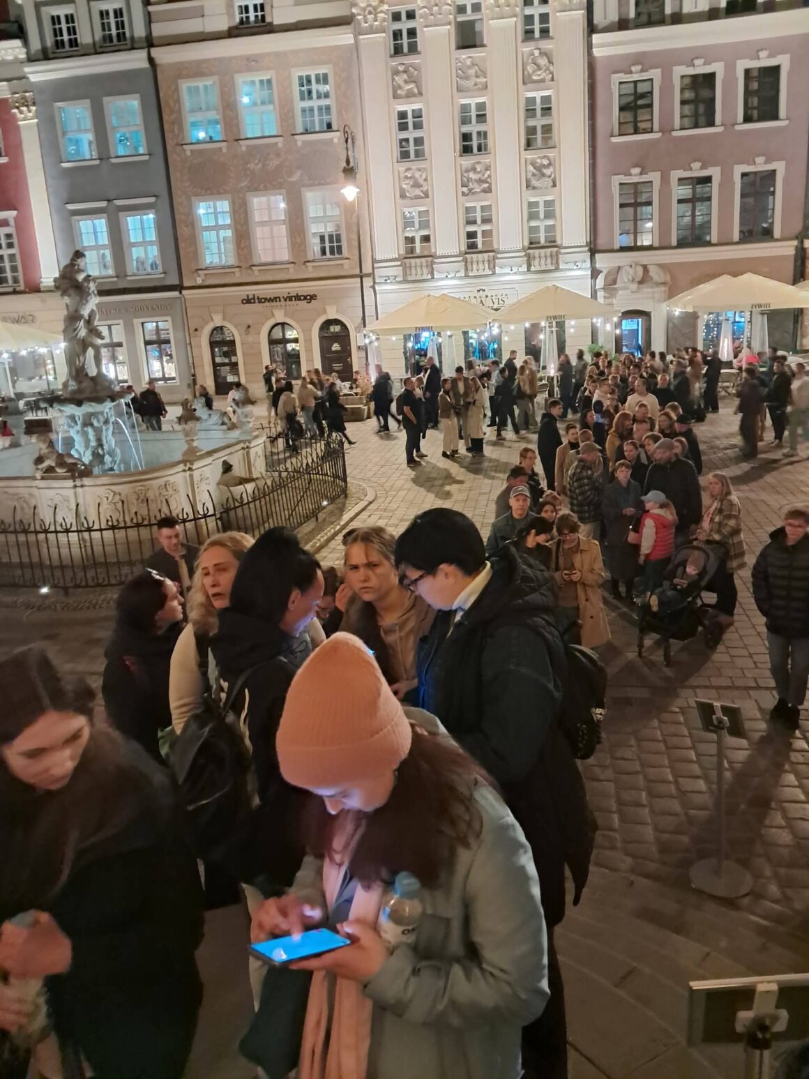 Fragment placu z kamienicami i fontanną. Ludzie stoją w długiej kolejce.