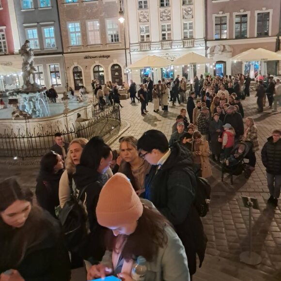 7 Fragment placu z kamienicami i fontanną. Ludzie stoją w długiej kolejce.