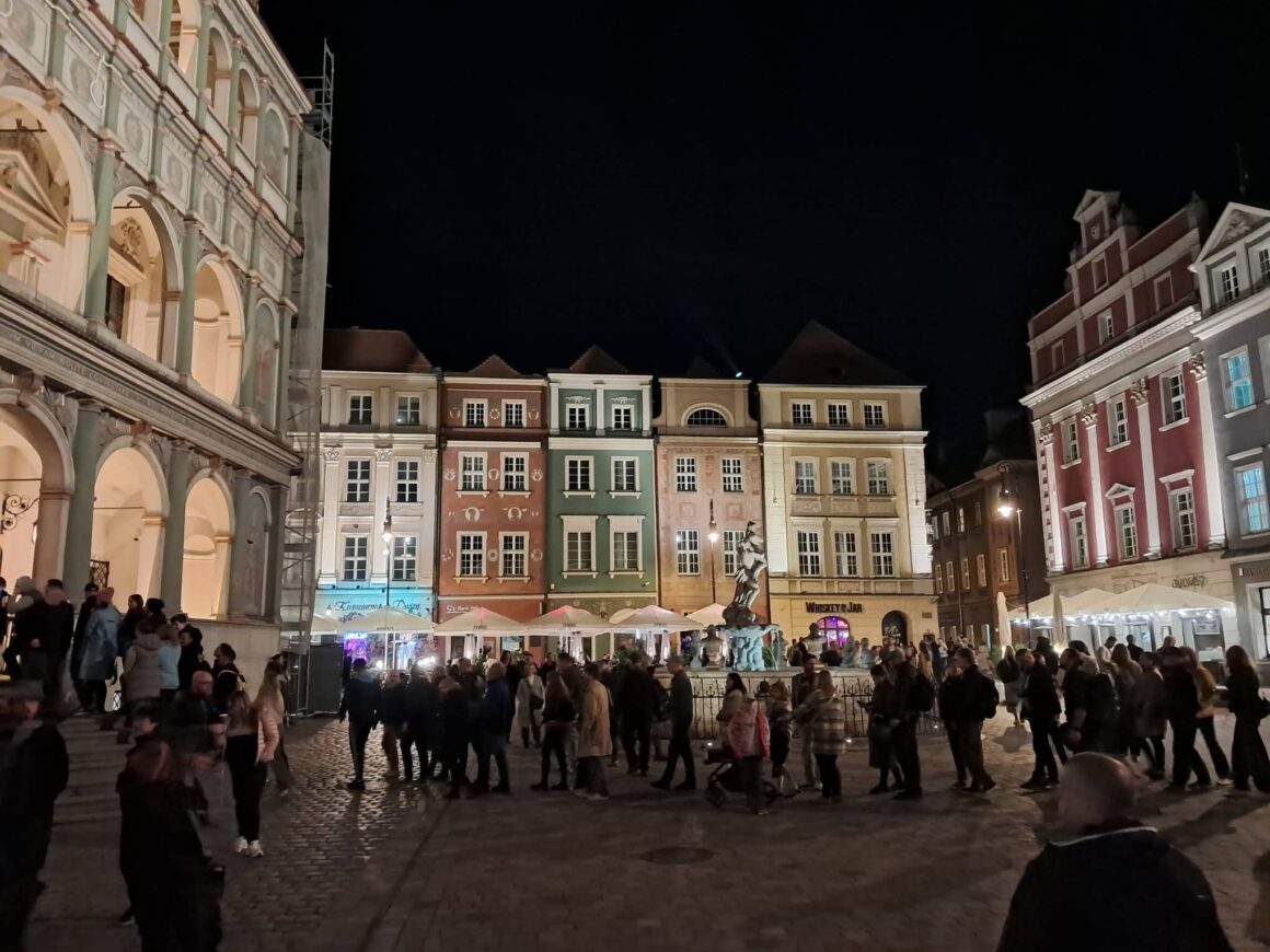 Fragment starego rynku w Poznaniu nocą. Na środku placu stoi wielu ludzi. Światło skierowane jest na elewacje kamienic.