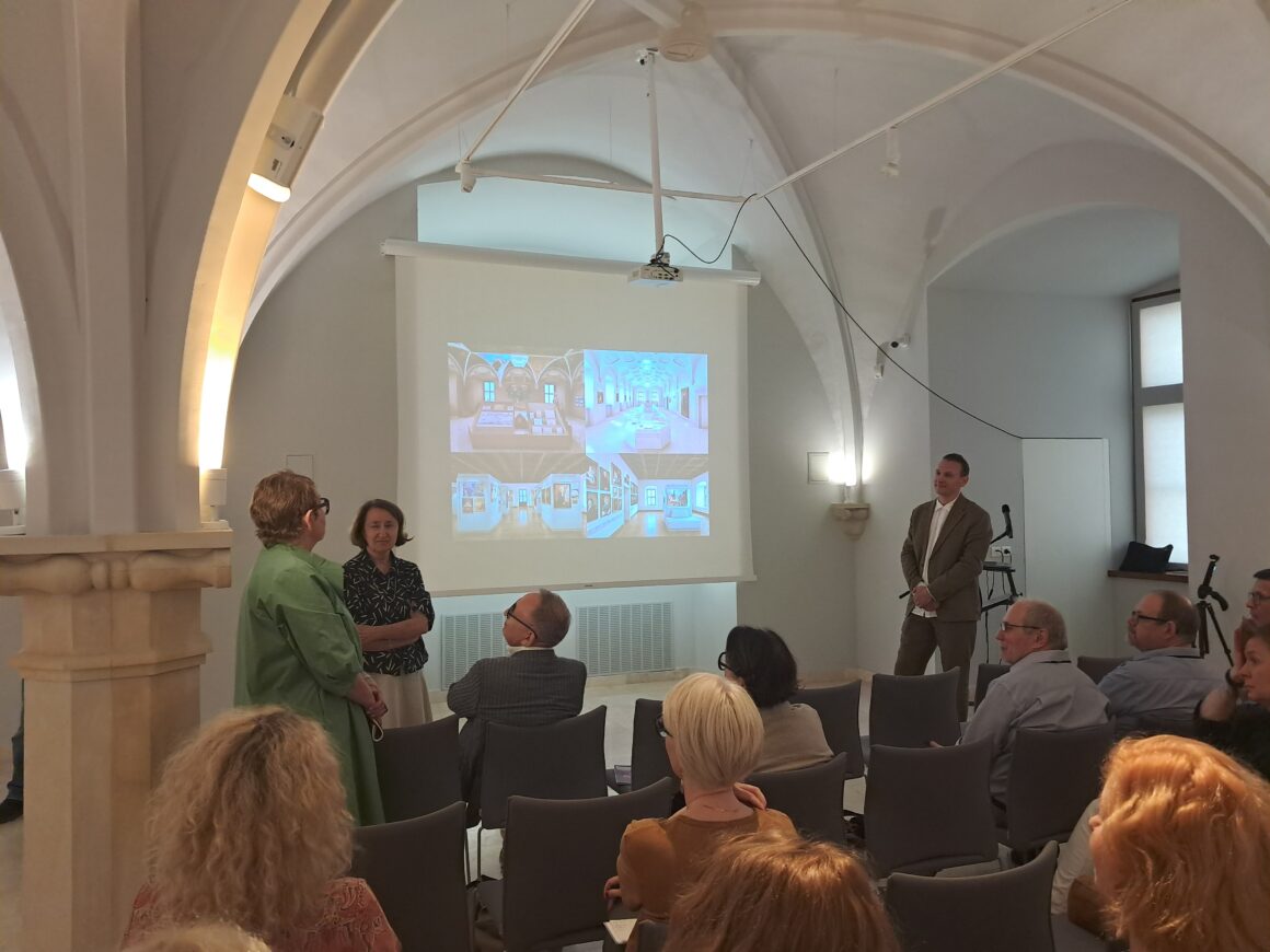 Zacieniona sala wykładowa. Po lewej stronie wyświetlony z rzutnika fragment prezentacji. Po prawej stroni, po lewej dwie kobiety. Na pierwszym planie siedzący na krzesłach słuchacze.