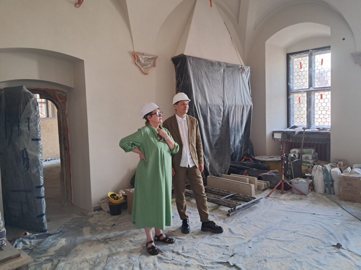 Kobieta i mężczyzna w białych kaskach budowlanych w zabytkowym pomieszczeniu. Na podłodze wyłożona folia ochronna. Przy oknie po prawej stornie leżą materiały budowlane.