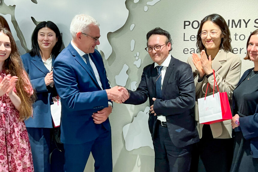 Na pierwszym planie dyrektor Muzeum Narodowego w Poznaniu ściska rękę dyrektorowi Voc Sou Kim z Korei. Wokół nich kilka osób z delegacji. Stoją na tle mapy świata.
