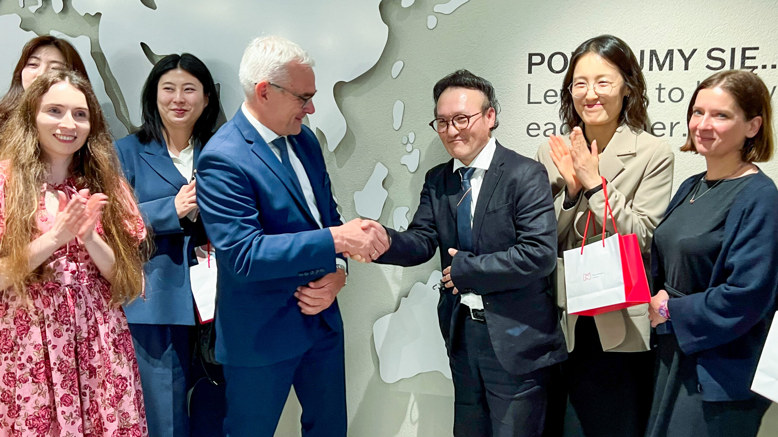 Na pierwszym planie dyrektor Muzeum Narodowego w Poznaniu ściska rękę dyrektorowi Voc Sou Kim z Korei. Wokół nich kilka osób z delegacji. Stoją na tle mapy świata.