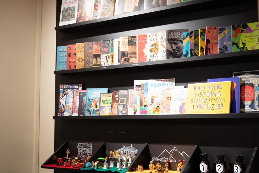 Czarny regał z książkami. Na najniższej półce ustawione są figurki Playmobile i flakony z zapachami.